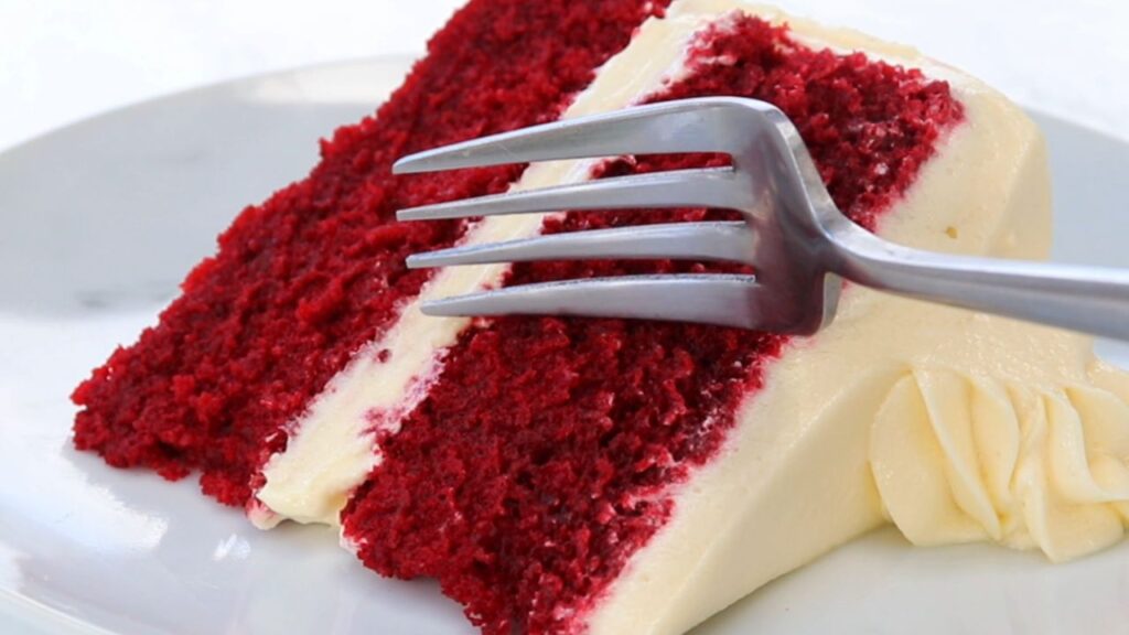Slice of red velvet cake with cream cheese frosting on a white plate, a silver fork resting against the cake edge.