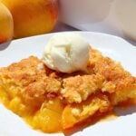 Slice of peach cobbler with a scoop of vanilla ice cream on a white plate, peaches visible in the background.