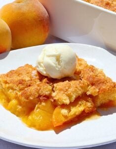 Slice of peach cobbler with a scoop of vanilla ice cream on a white plate, peaches visible in the background.