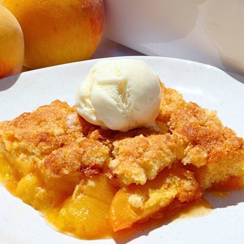 Slice of peach cobbler with a scoop of vanilla ice cream on a white plate, peaches visible in the background.