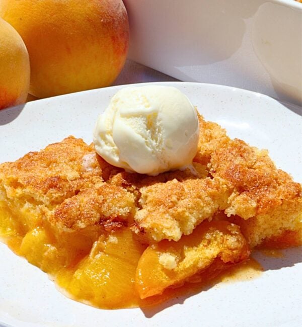 Slice of peach cobbler with a scoop of vanilla ice cream on a white plate, peaches visible in the background.