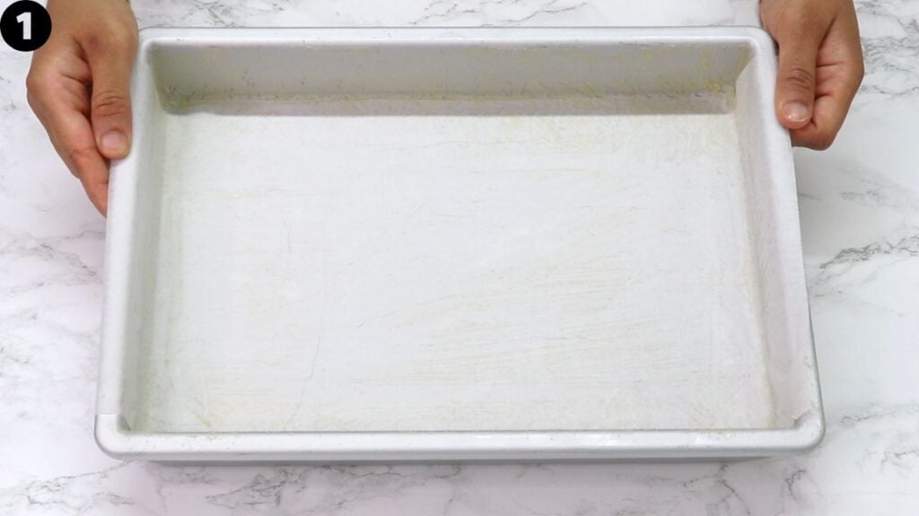 Hands hold a parchment-white baking tray over a marble countertop, empty and clean with slight stains around edges.