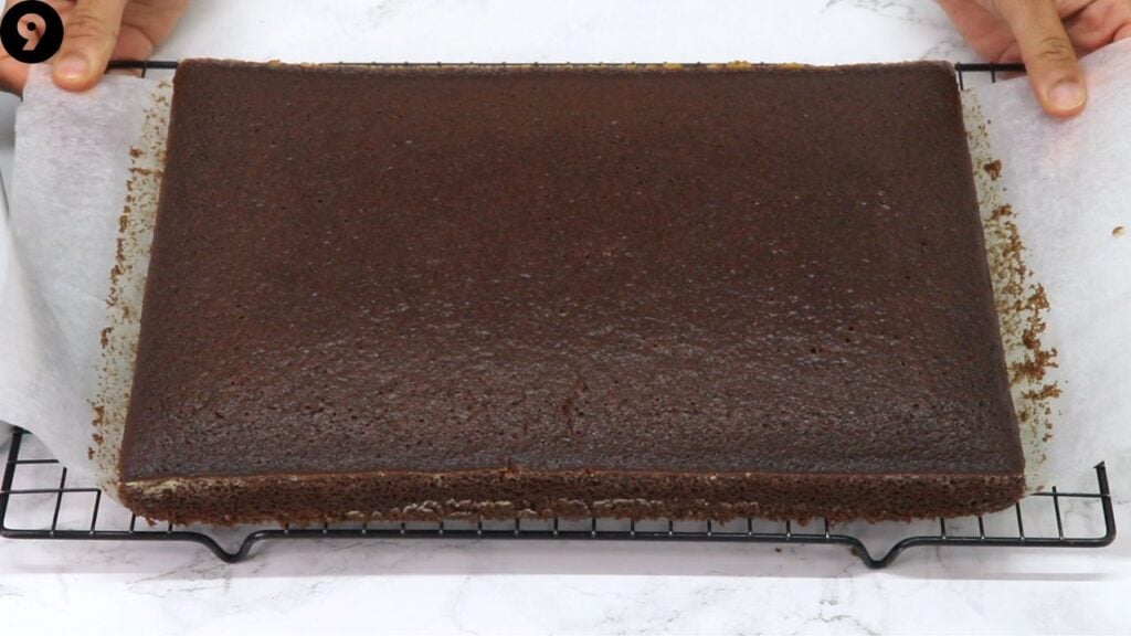 Large rectangular chocolate cake cooling on a wire rack with parchment paper.