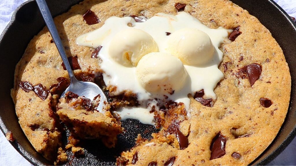 pizookie - Cakes by MK Warm chocolate chip cookie served in a cast‑iron skillet with three scoops of vanilla ice cream and a spoon, a bite taken.