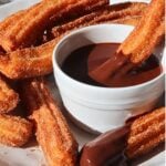 Crispy cinnamon-sugar churros arranged around a white bowl of melted chocolate for dipping.