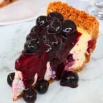 Slice of vanilla cake with a glossy blueberry cherry sauce dripping over it and a crumbly golden crust on the edge, served on a white plate