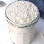 Close-up of a glass jar filled to the top with white powder (likely flour), the surface slightly uneven and floury.
