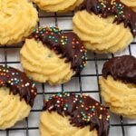 Ruffled vanilla cookies half-dipped in chocolate with colorful sprinkles on a cooling rack.