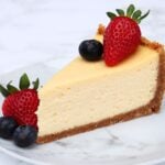Slice of creamy cheesecake on a graham crust, topped with a strawberry and blueberries on a marble plate.