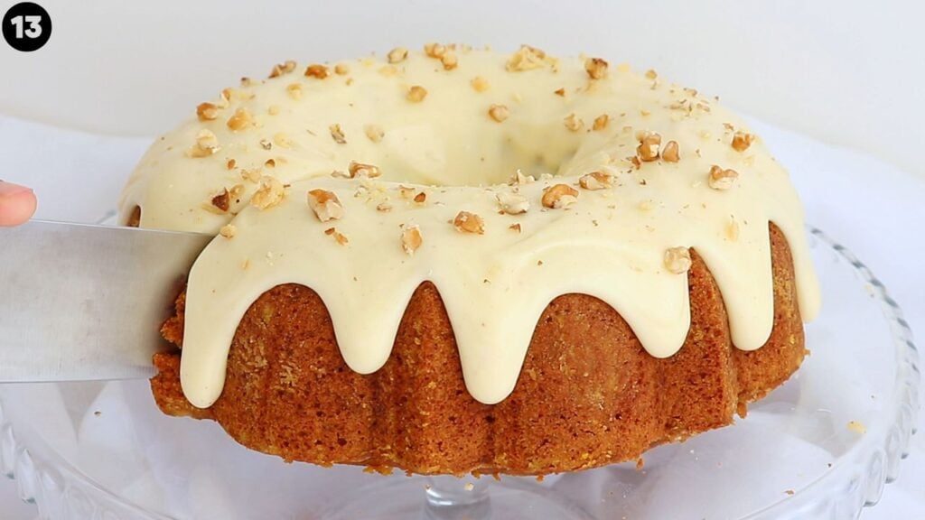 pumpkin bundt cake - Cakes by MK Bundt cake with thick white glaze and chopped nuts on top, being sliced with a knife on a glass cake stand