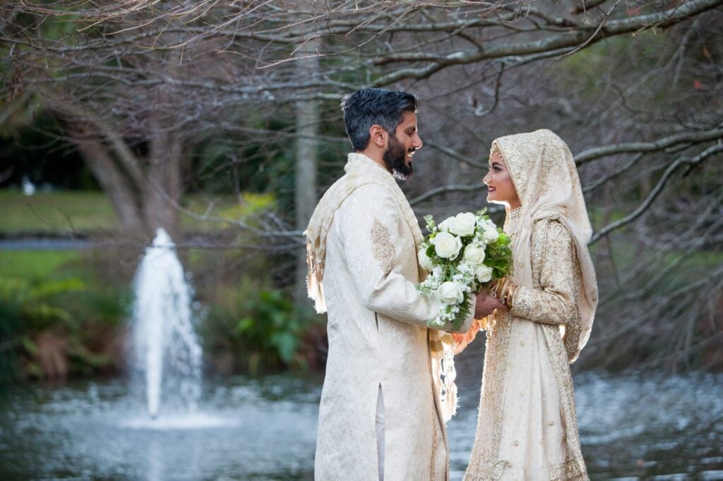 Bride and groom in cream-gold traditional attire exchange smiles by a lake with a fountain in the background, holding a bouquet of white roses.