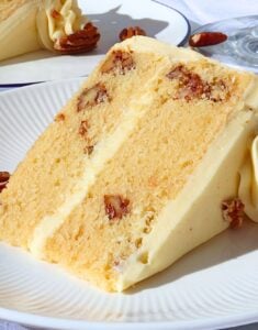 Slice of vanilla cake with creamy frosting and pecan pieces embedded, on a white plate with decorative frosting in the background.