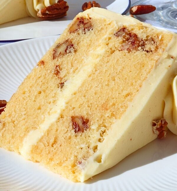Slice of vanilla cake with creamy frosting and pecan pieces embedded, on a white plate with decorative frosting in the background.