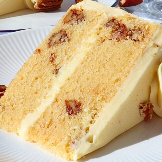Slice of vanilla cake with creamy frosting and pecan pieces embedded, on a white plate with decorative frosting in the background.