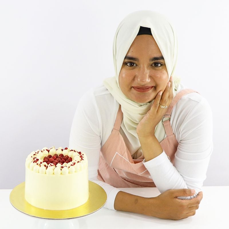 Woman in a light hijab and pink apron sits at a white table with a cream cake topped with berries, smiling gently.