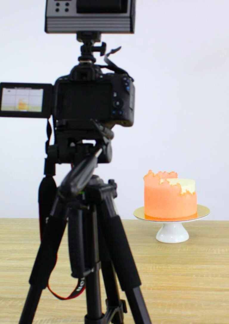 Camera on a tripod filming an orange frosted cake on a white stand in a studio setting