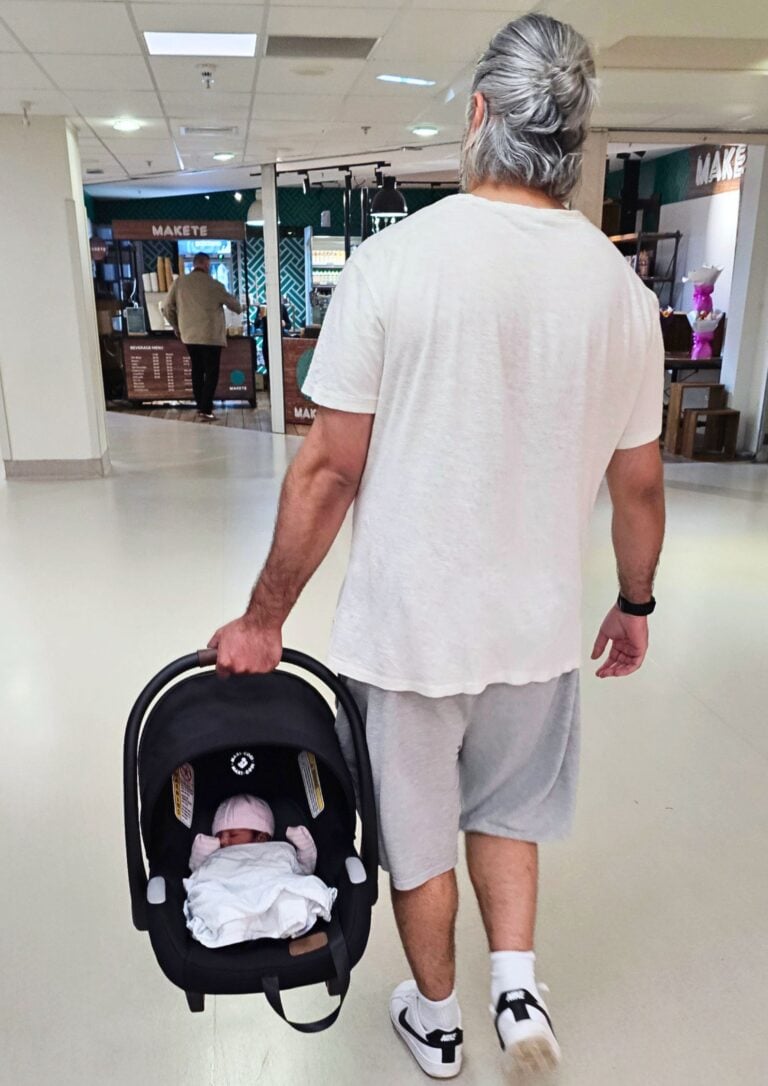 Adult with gray hair pushing an infant in a black infant car seat stroller through a shopping mall kiosk area.