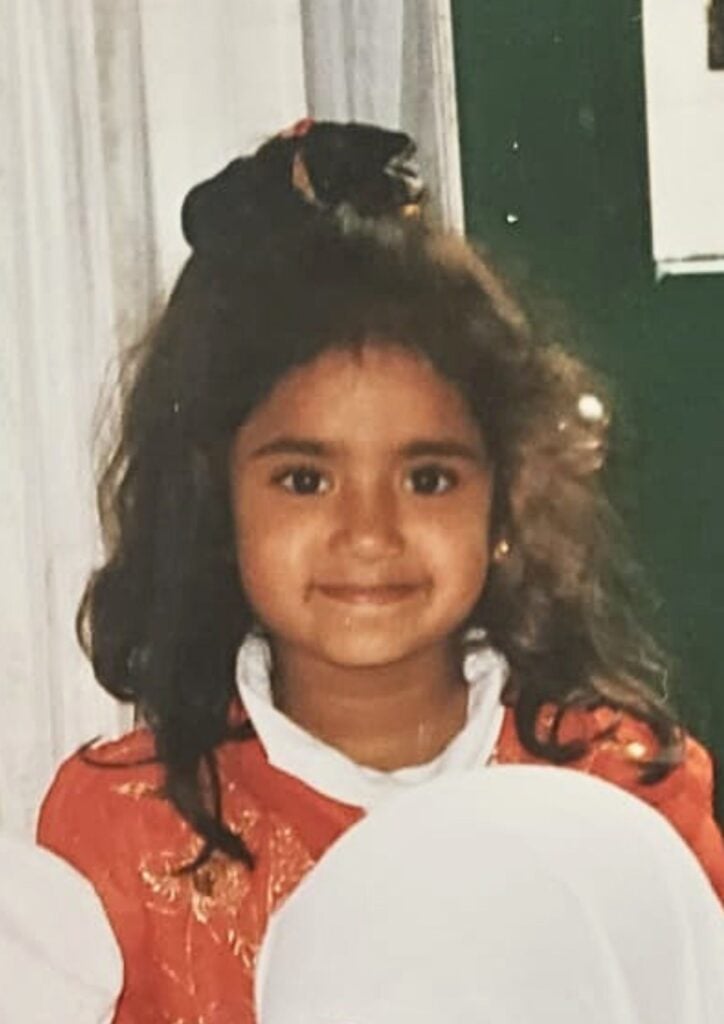 Close-up of a young girl with dark hair wearing a red dress with a white collar, smiling at the camera.
