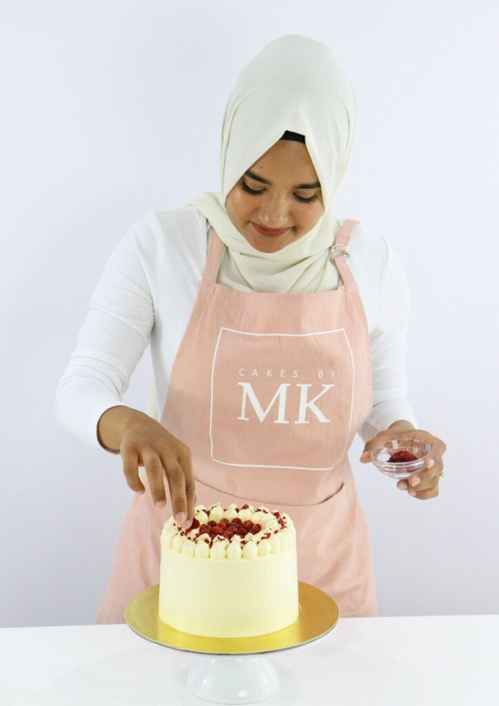 Woman wearing a cream hijab and pink apron decorates a yellow-frosted cake with berries on top.